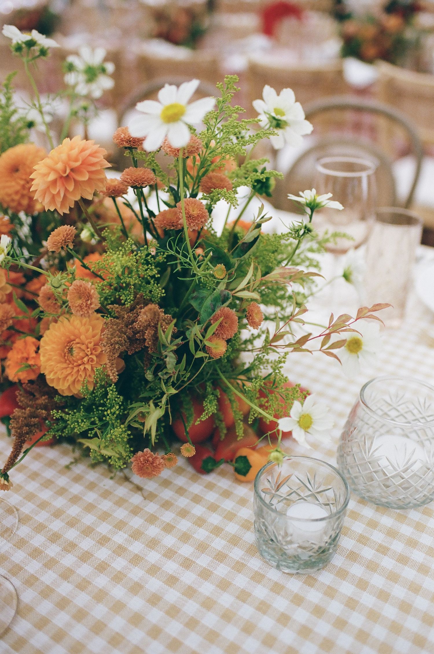 Orange dahlias and white daisies centerpiece on gingham tablecloth with crystal glassware at elegant event.