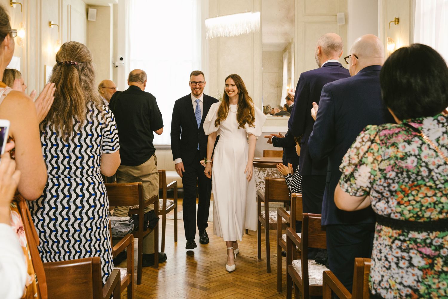Bride in white dress walks down aisle with escort at elegant indoor wedding ceremony with guests watching.