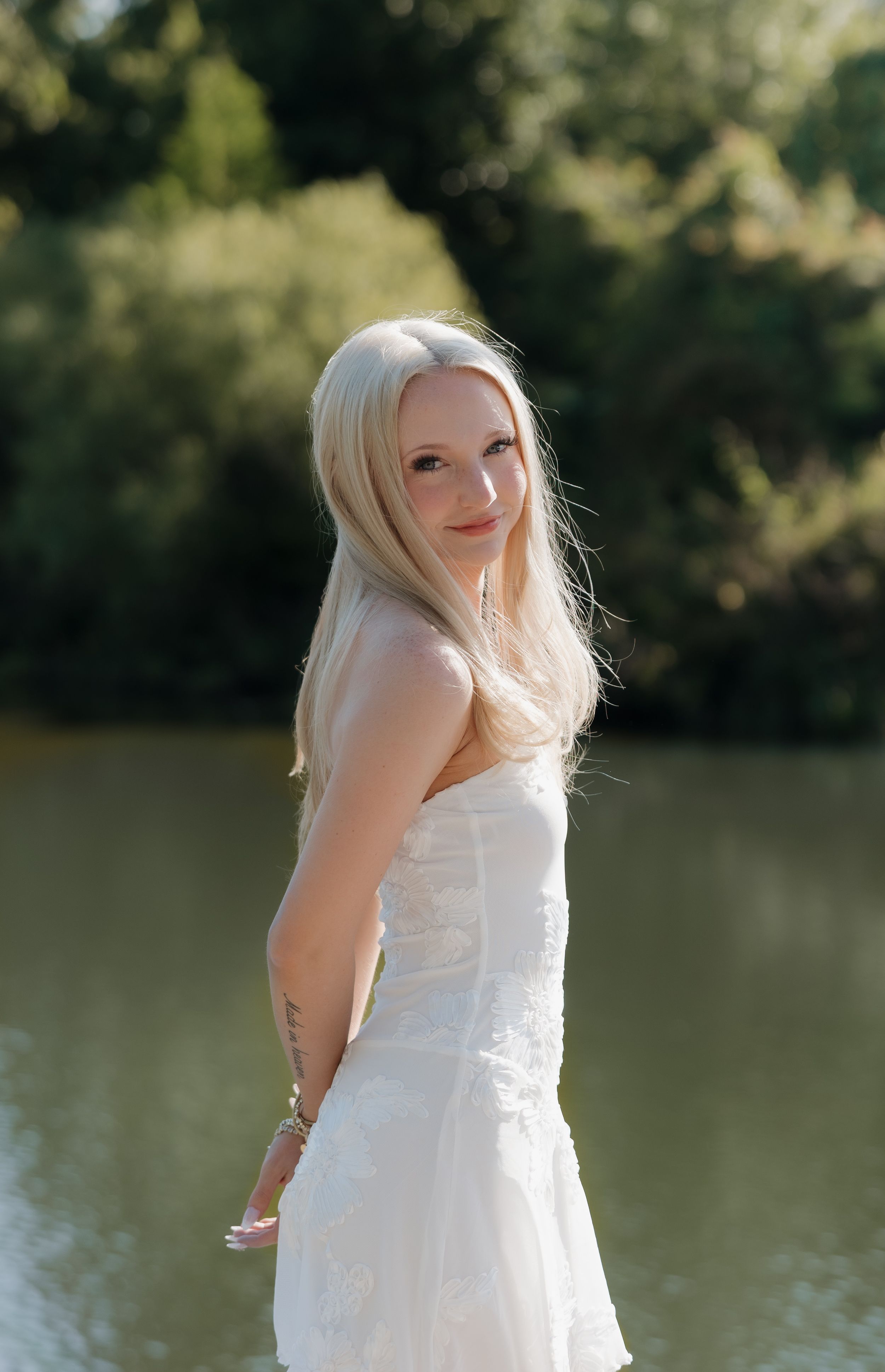 Blonde woman in white strapless dress standing near water with green trees in background.
