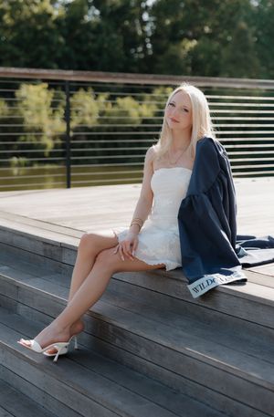 Blonde woman in white dress and heels sitting on wooden deck steps with navy graduation gown beside her.