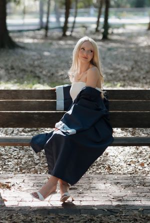 Blonde woman in navy graduation gown sits gracefully on a park bench, smiling over her shoulder in dappled sunlight.