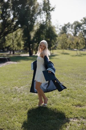 Blonde woman in white mini dress and navy coat posing outdoors in sunny green park.