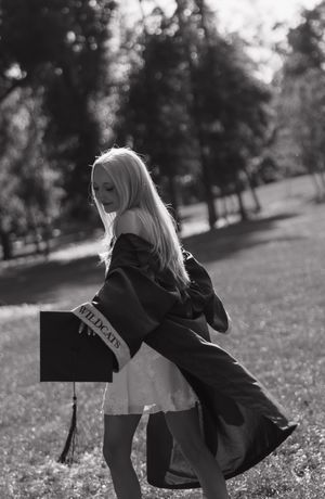 Black and white photo of a blonde graduate in cap and gown outdoors, sash reading Wildcats, posing on grass.
