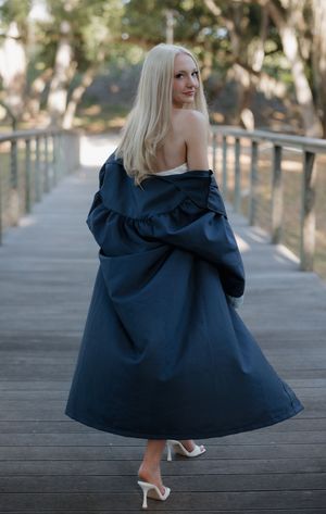 Blonde woman in elegant navy off-shoulder gown on wooden bridge, viewed from behind.