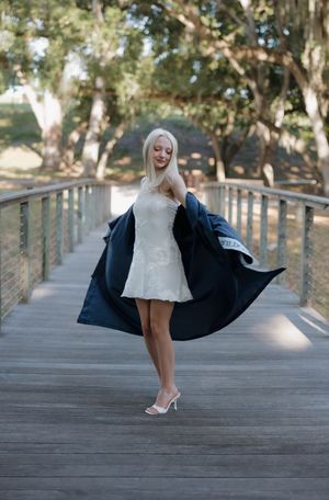 Blonde woman in white lace dress and navy coat posing on wooden bridge surrounded by trees.