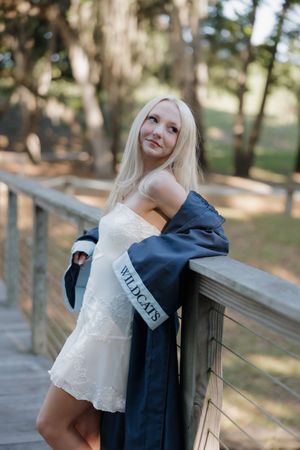 Blonde woman in white dress and navy Wildcats graduation gown leaning on wooden fence in park.