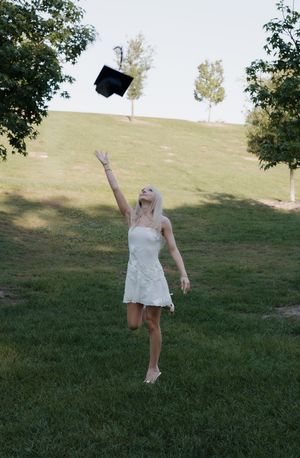 Blonde woman in white dress tossing graduation cap in the air outdoors.