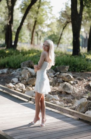 Blonde woman in white strapless mini dress and heels poses on wooden boardwalk surrounded by rocks and trees.