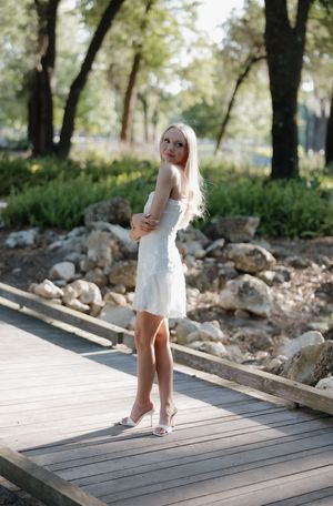 Blonde woman in white mini dress and heels standing on wooden boardwalk surrounded by rocks and trees.