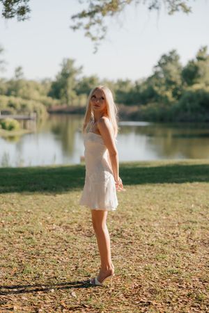 Blonde woman in white sundress standing barefoot on grass near a serene lake in a sunny park.