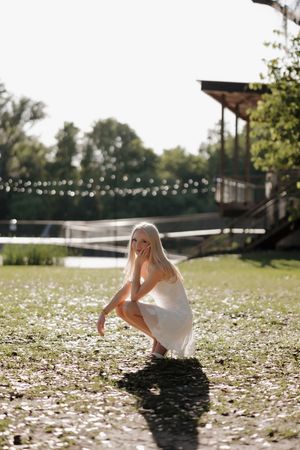 Blonde woman in white dress crouching on flower-covered ground outdoors.