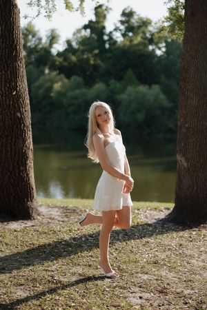 Blonde woman in white dress posing barefoot between trees near a lake.