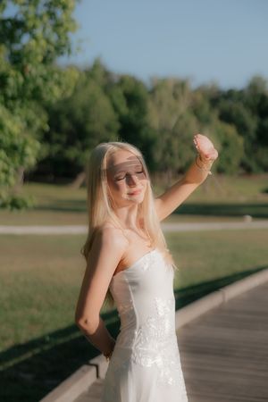 Young blonde woman in white lace dress posing gracefully outdoors in a sunny green park.