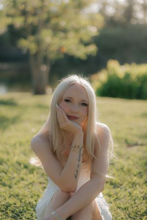 Young blonde woman sitting on grass in golden sunlight wearing white dress with bracelet tattoo.