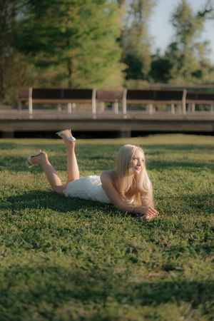 Smiling blonde woman in white dress lying on green grass in sunny park.