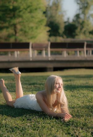 Smiling blonde woman in white dress lying on green grass in sunny park with wooden bridge background.