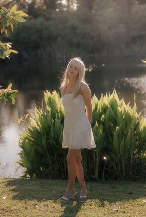 Blonde woman in white sundress standing by lush green plants near a sunlit pond.