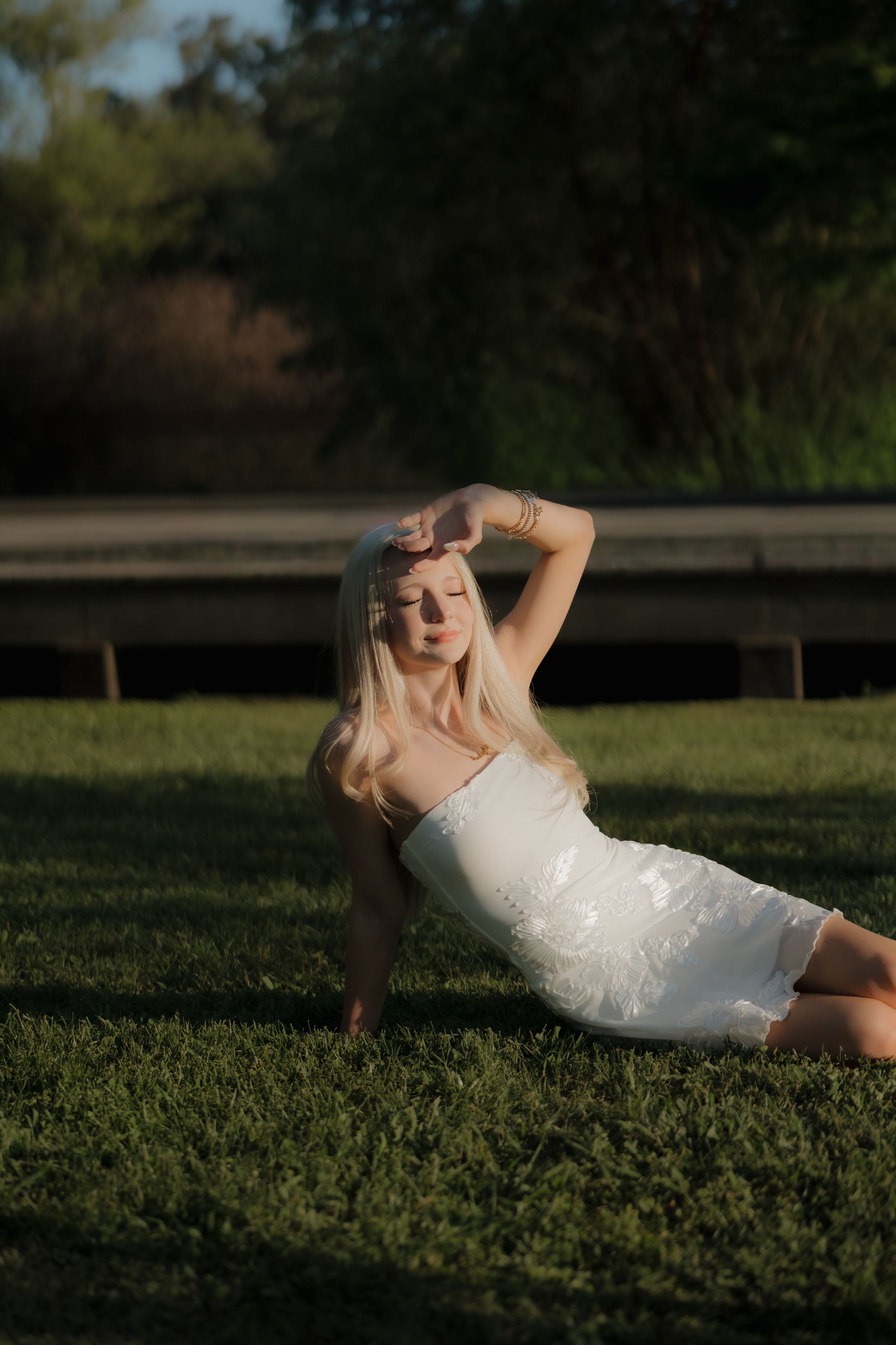 Blonde woman in white dress posing on green grass in golden hour sunlight with trees in background.