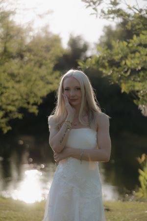 Blonde woman in white strapless dress standing near pond in sunlit garden with hand on cheek.
