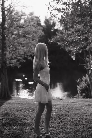 Black and white photo of a blonde woman in a white dress standing by a reflective pond surrounded by lush trees.