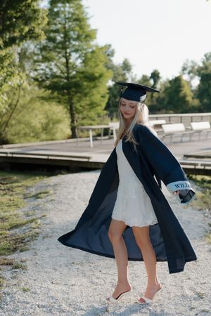 Blonde woman in graduation cap and gown over white dress, walking barefoot outdoors near a dock.