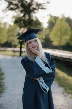 Blonde graduate in cap and gown posing outdoors with Wildcats sash.