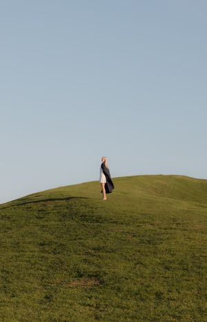 Woman in white dress and graduation gown standing atop a green hill.