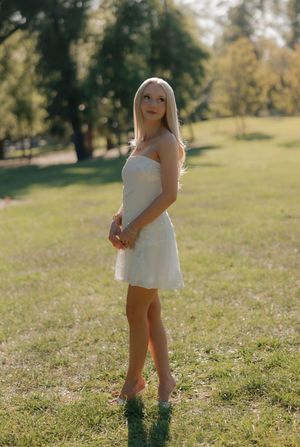 Blonde woman in white strapless mini dress standing barefoot on grass in sunny park.