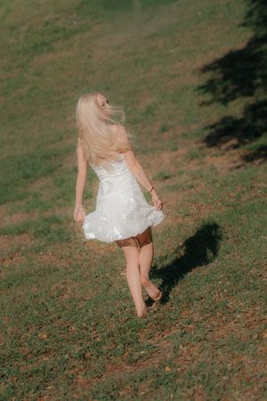 Blonde woman in white dress walking barefoot across green grassy field.