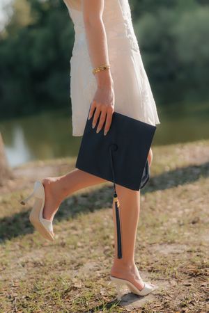 Close-up of a graduate in a white dress holding a black graduation cap while wearing gold bracelet and heels.