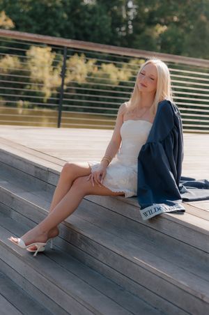 Blonde woman in white dress sitting on wooden steps with graduation gown.