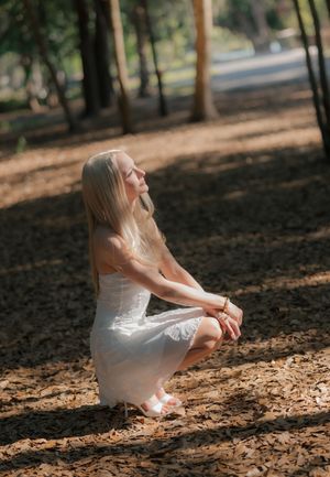 Blonde woman in white dress crouching peacefully in sunlit pine forest.