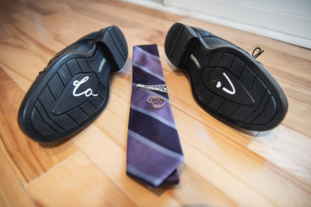 Black dress shoes with soles up, purple striped tie, and wedding rings arranged on hardwood floor.