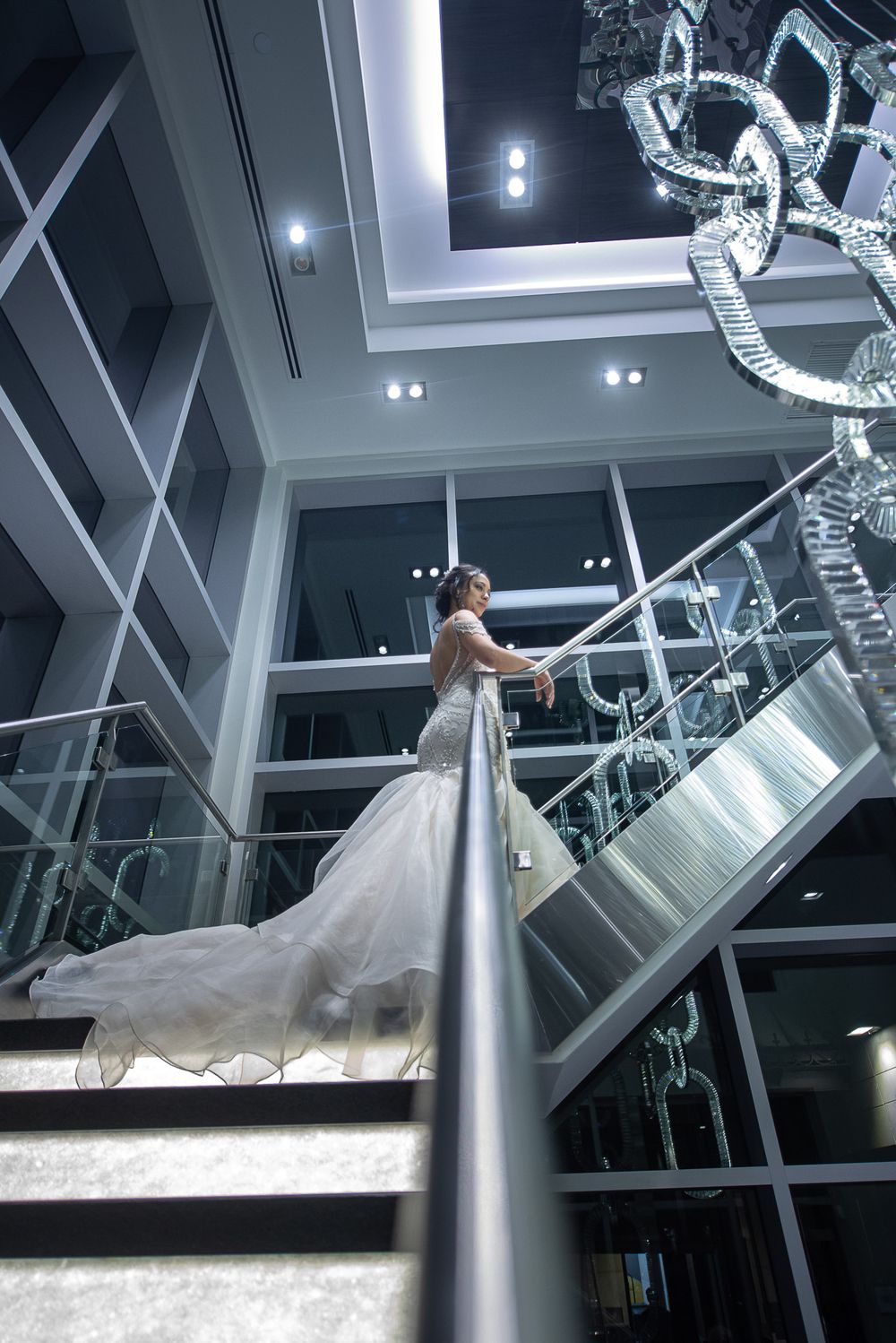 Bride in mermaid wedding dress with flowing train standing on modern staircase with crystal chandelier.