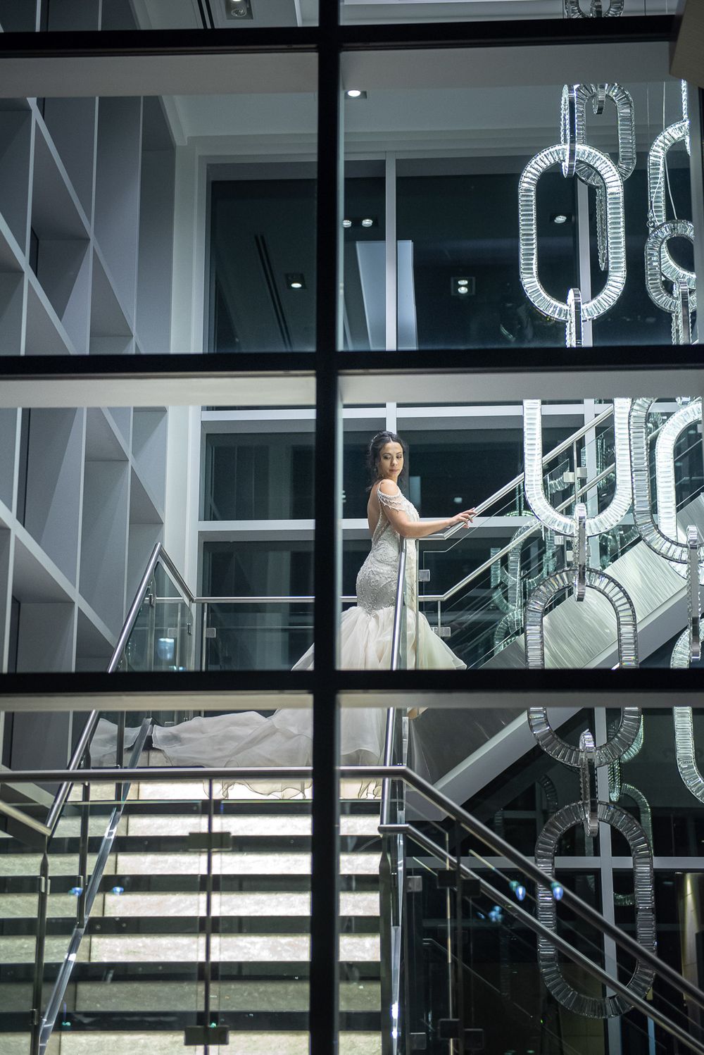 Bride in fitted white wedding gown posing on staircase viewed through glass wall with chain sculpture.