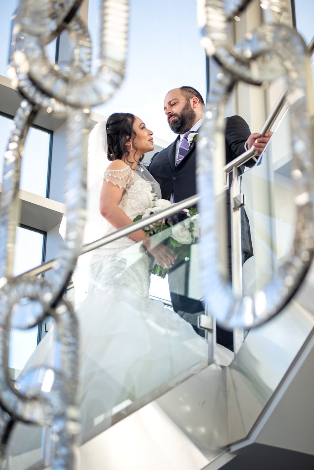 Bride and groom share an intimate moment on a glass staircase framed by crystal chandelier details.