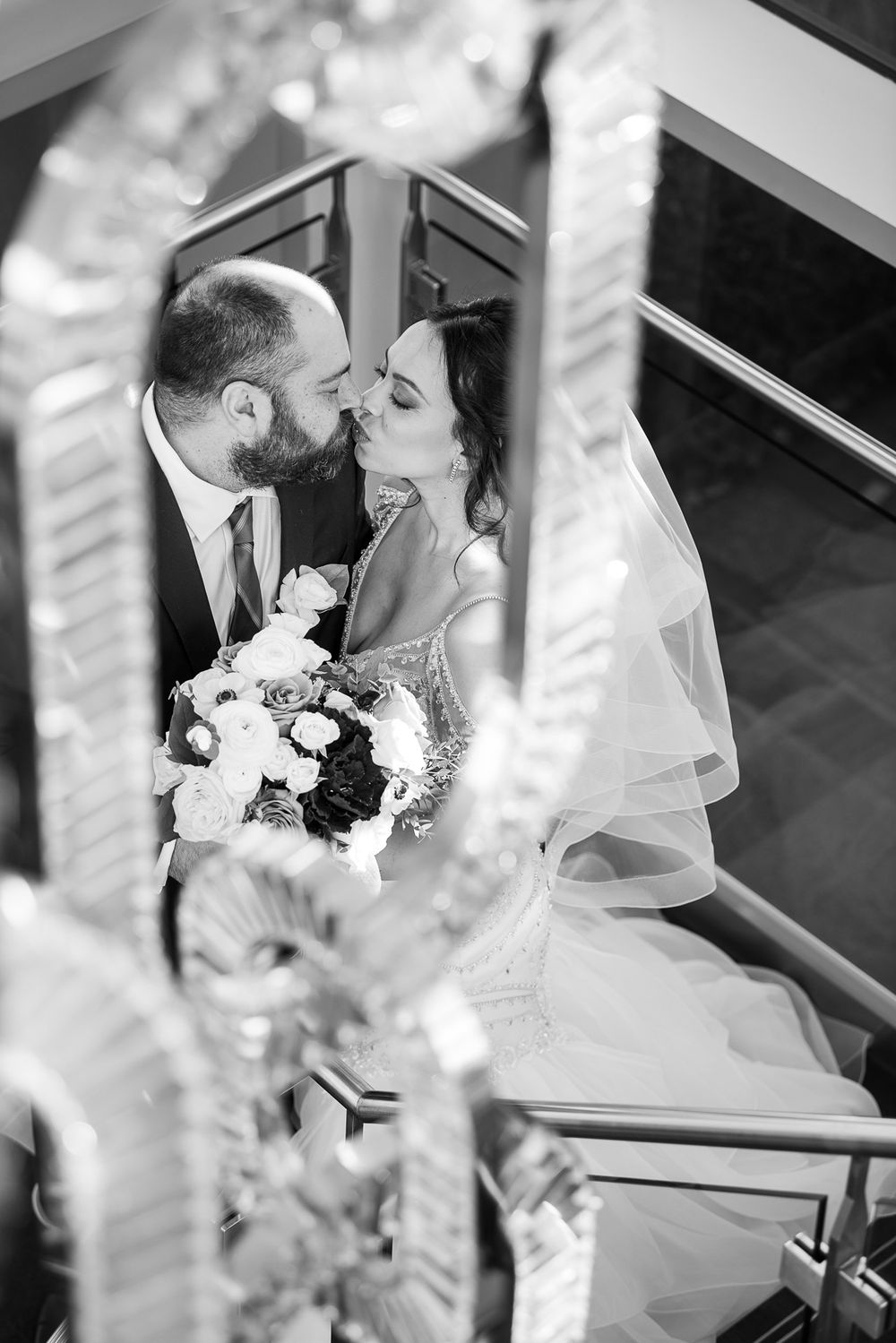 Black and white photo of bride and groom sharing a kiss on a staircase with chandelier in foreground.