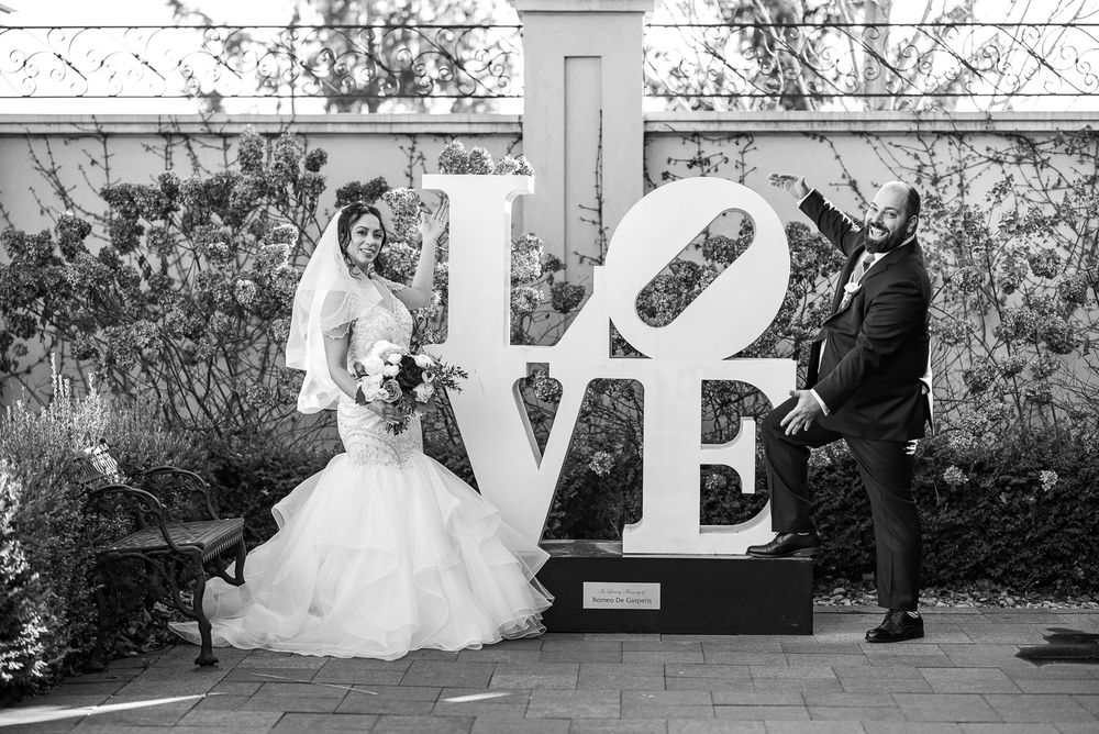 Black and white photo of bride and groom posing playfully beside a large LOVE sculpture outdoors.