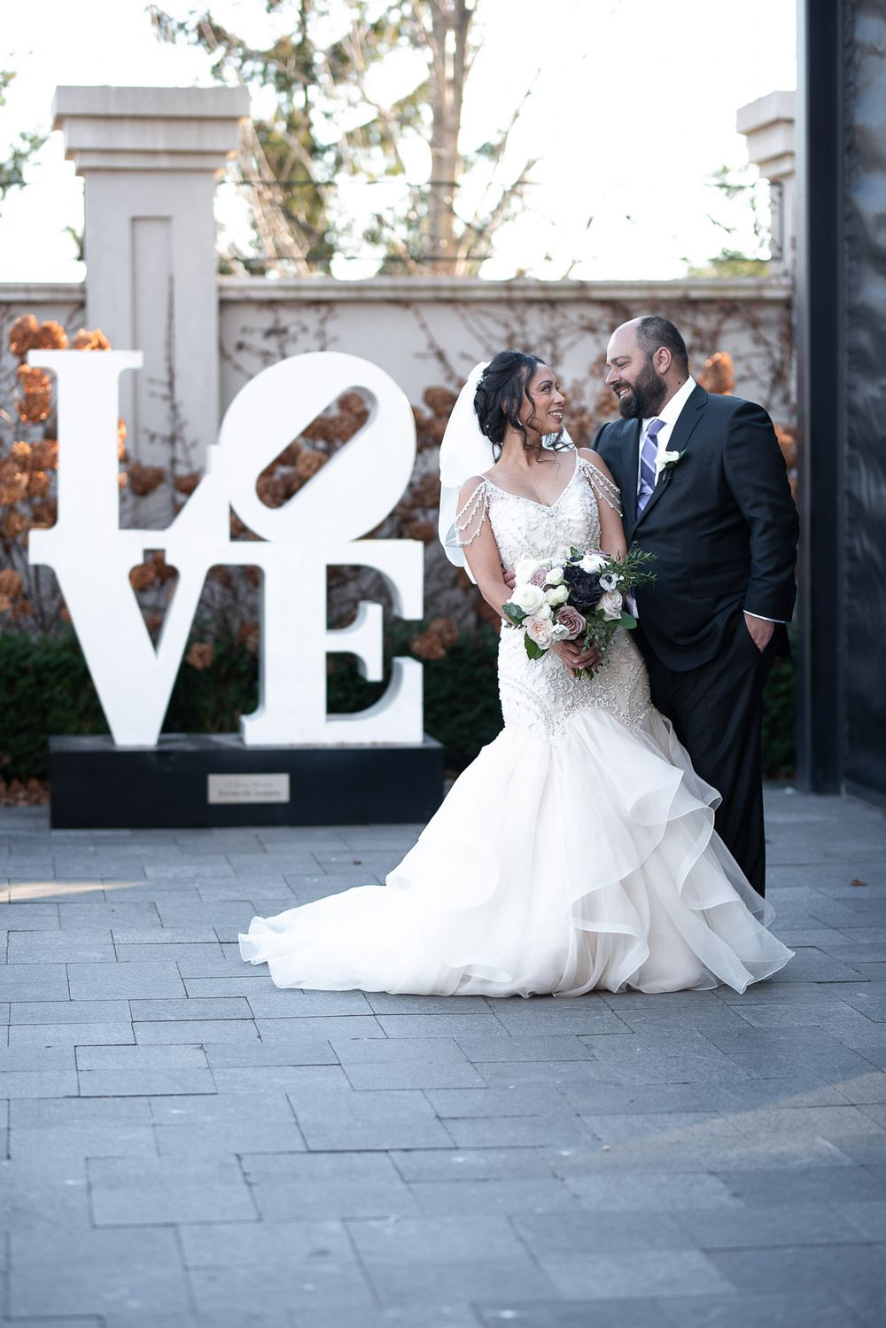 Bride and groom share intimate moment in front of large white LOVE sculpture on stone patio.