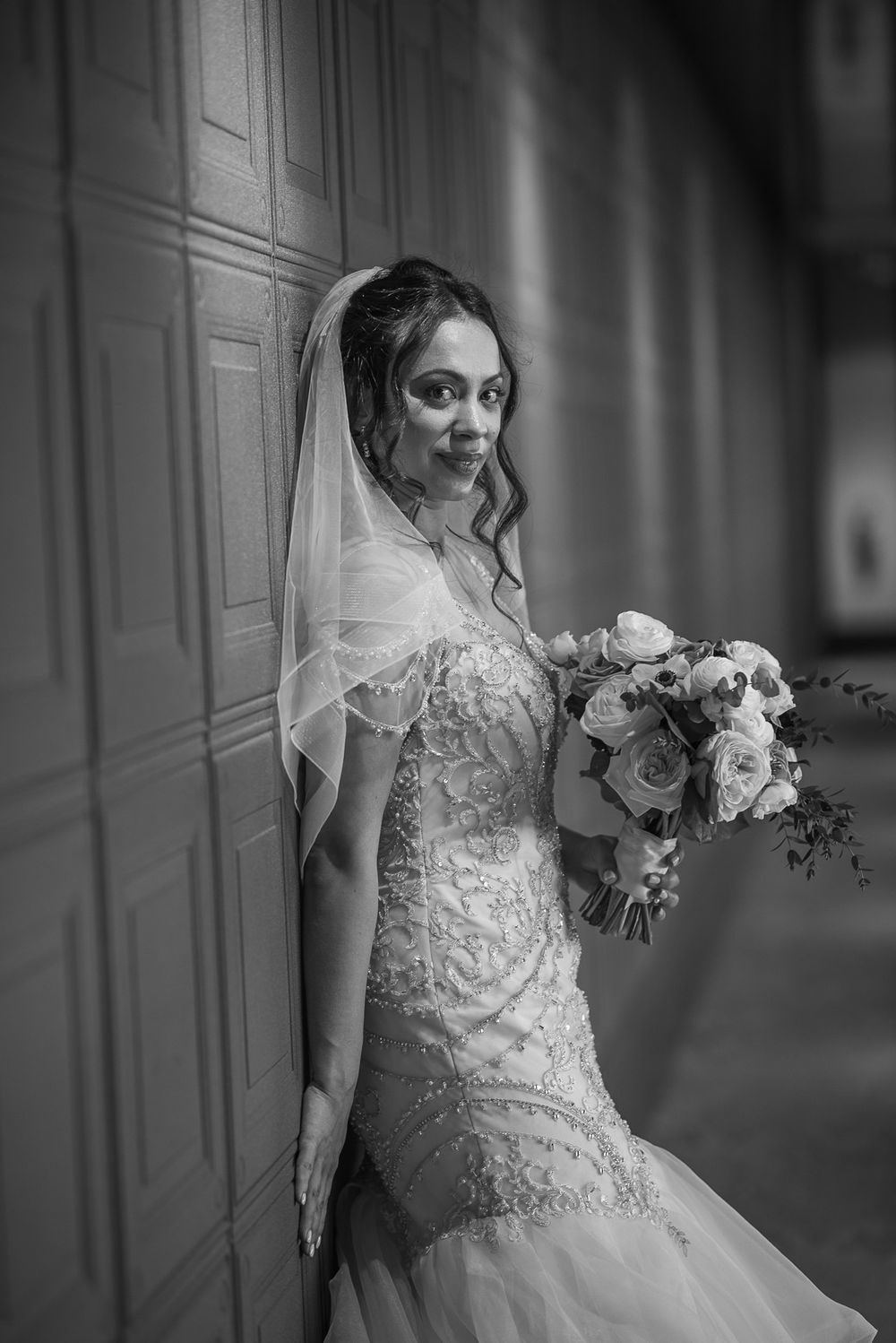 Black and white portrait of a bride in a lace gown leaning against a wooden door.