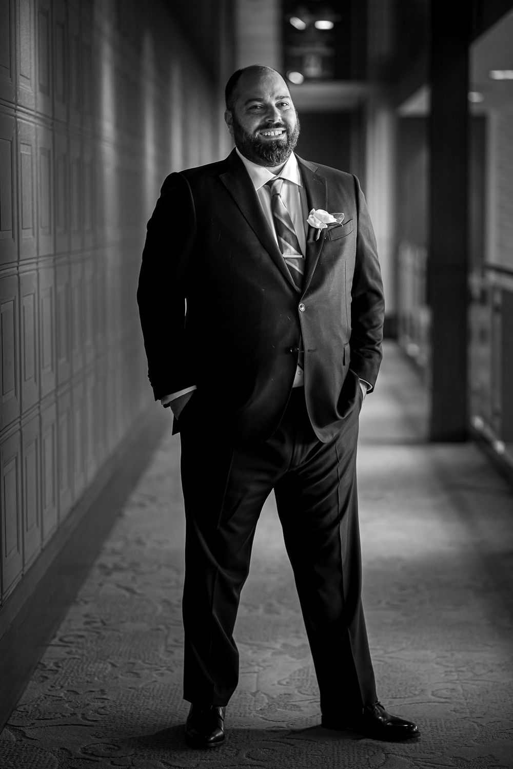 Black and white portrait of a smiling groom in a dark suit standing in a hotel corridor.