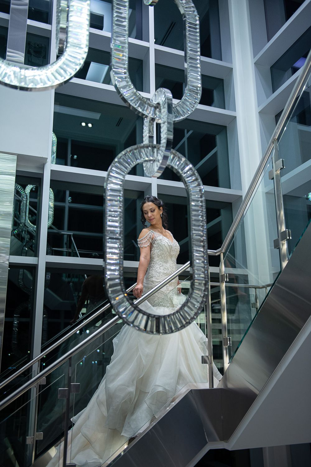 Bride in lace mermaid wedding dress posing on staircase framed by large crystal chain sculpture.