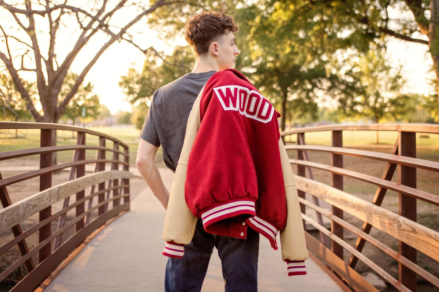 Senior boy holding red varsity jacket on a wooden bridge at golden hour at Bear Creek Park in Keller, TX.