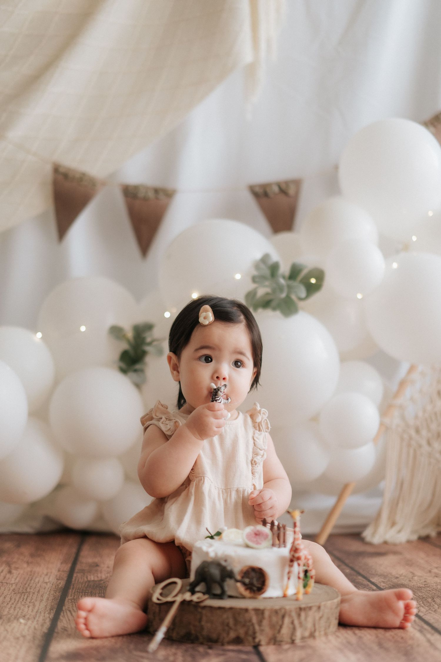 Baby in pink dress sits on floor with white balloons and bunting decorations for first birthday celebration.