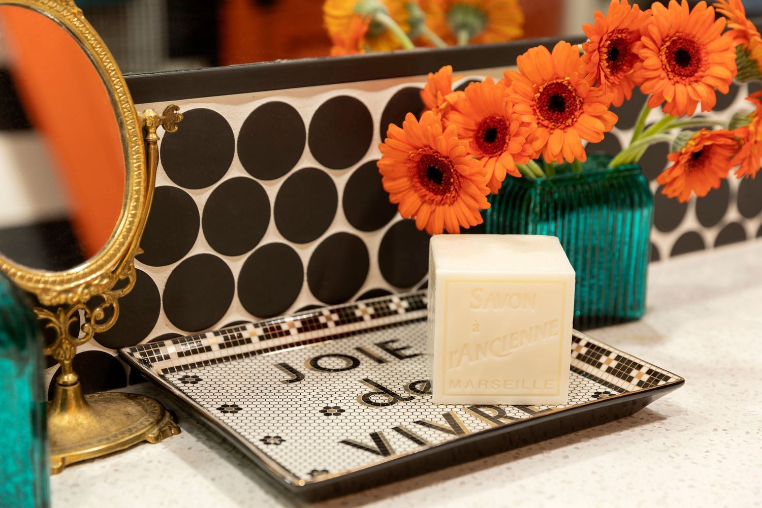 Orange gerbera daisies sit beside a black and white patterned tray with a gold-rimmed mirror and teal vase.