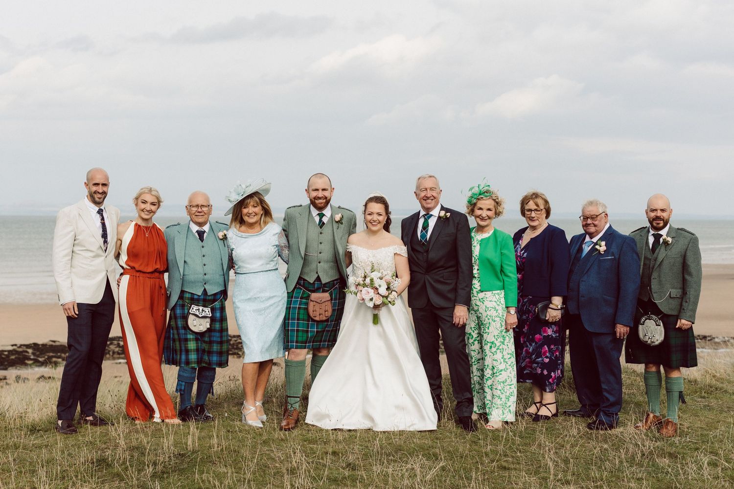 Wedding party celebrates on a scenic beach with guests wearing colorful formal attire and traditional Scottish kilts.