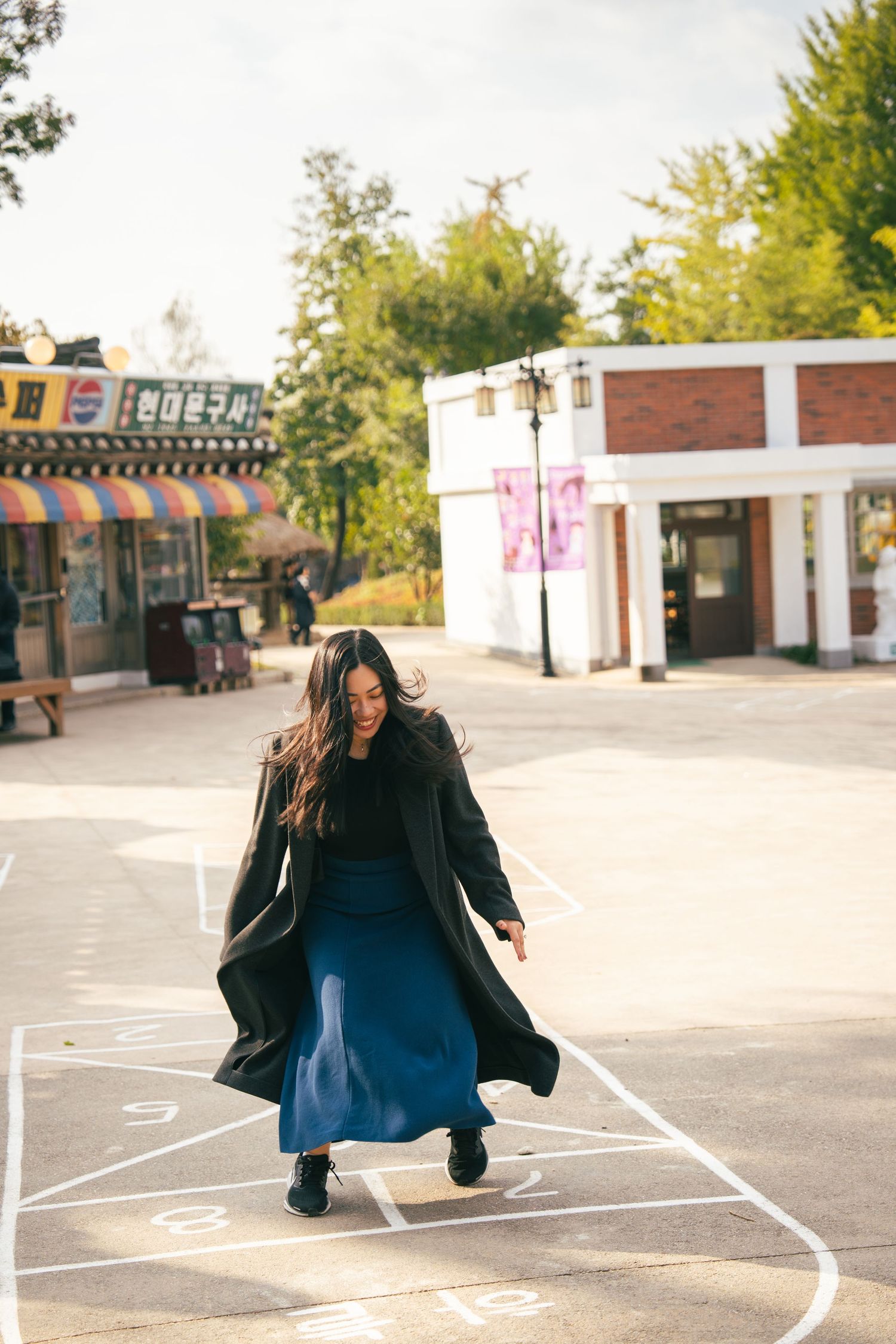 A person in a flowing black coat and blue skirt plays hopscotch on a sidewalk near storefronts on a sunny day.