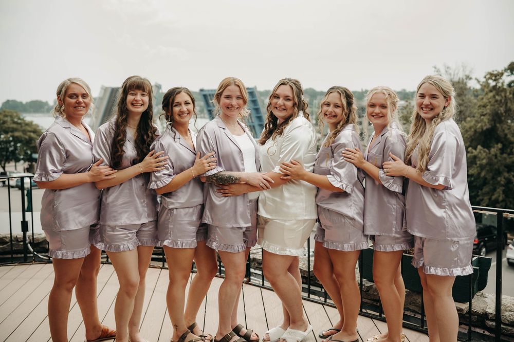 Bride and bridesmaids smile together in matching silver satin pajamas on an outdoor balcony before the wedding.
