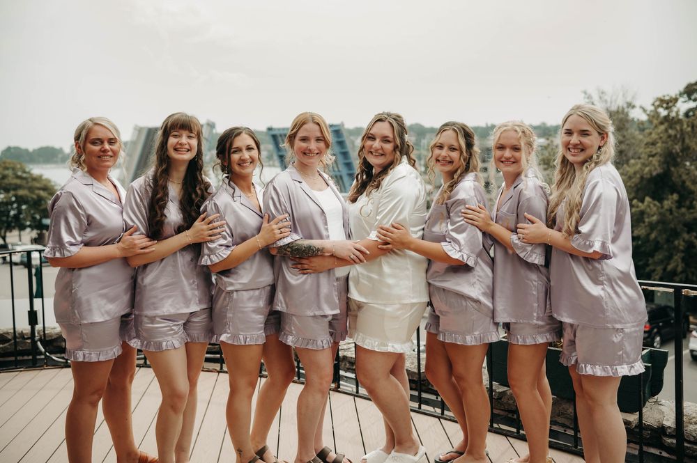 Bride in white robe poses with seven bridesmaids in matching silver satin pajama sets on a rooftop balcony.