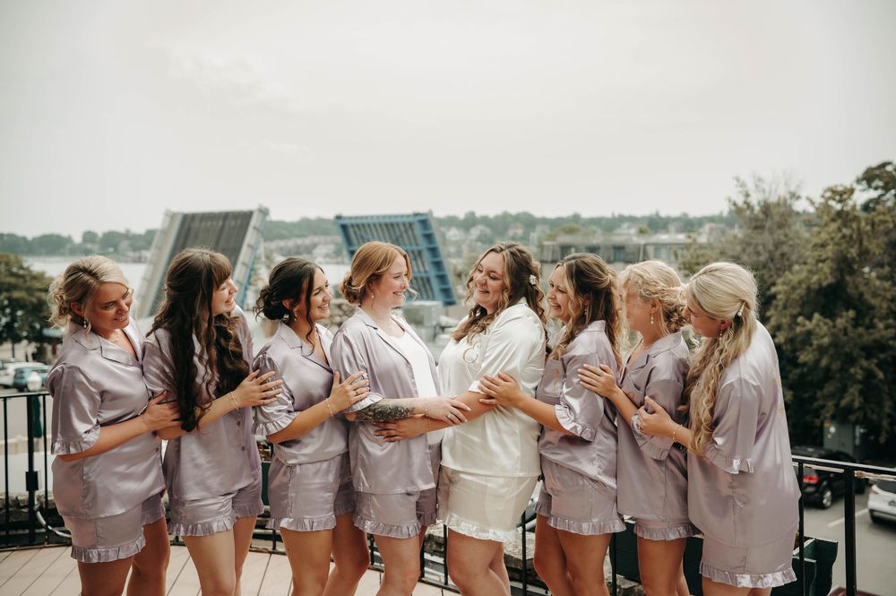 Bride and bridesmaids laugh together in matching satin pajamas on a rooftop balcony on the wedding morning.